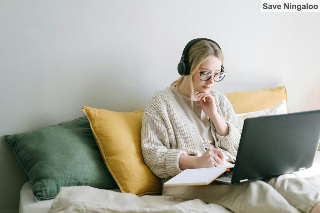 A focused woman in glasses and headphones works on a laptop from a cozy bed.