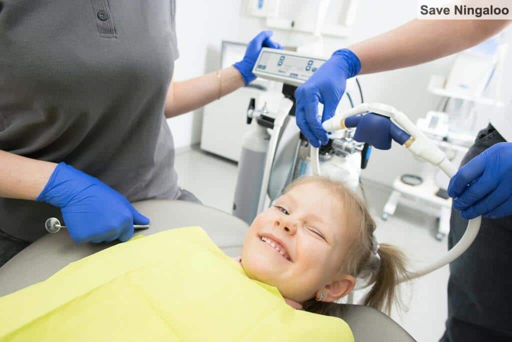 A cheerful child winks while sitting in a dental chair, surrounded by dental professionals.